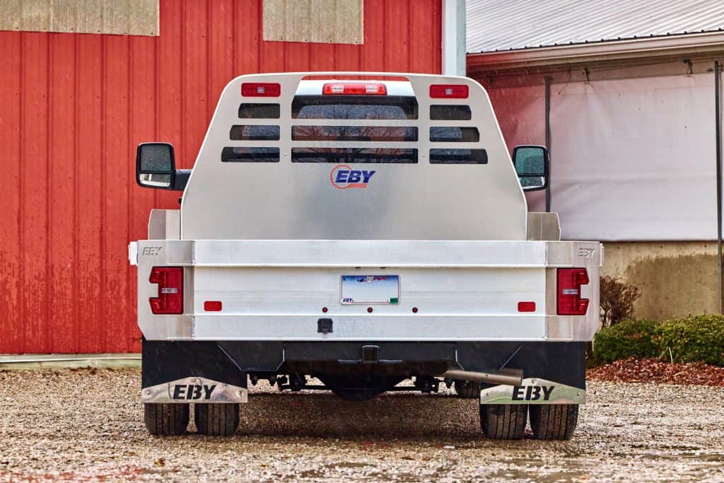 Rear view of a truck equipped with an EBY Big Country aluminum flat bed, featuring a slatted headache rack with the EBY logo, aluminum side rails, integrated tail lights, trailer hitch, and EBY-branded mud flaps, parked on gravel in front of a red barn.