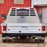 Rear view of a truck equipped with an EBY Big Country aluminum flat bed, featuring a slatted headache rack with the EBY logo, aluminum side rails, integrated tail lights, trailer hitch, and EBY-branded mud flaps, parked on gravel in front of a red barn.