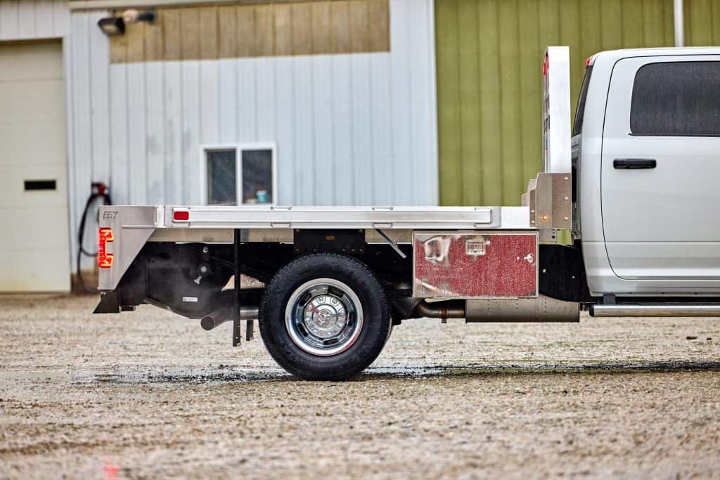 Side profile view of an EBY Big Country aluminum flat bed mounted on a white heavy-duty truck, showing the open flat deck, rear tail lights, EBY-branded side rail, chrome dual rear wheels, and a red under-bed storage box, parked on gravel in front of a metal farm building.