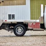 Side profile view of an EBY Big Country aluminum flat bed mounted on a white heavy-duty truck, showing the open flat deck, rear tail lights, EBY-branded side rail, chrome dual rear wheels, and a red under-bed storage box, parked on gravel in front of a metal farm building.