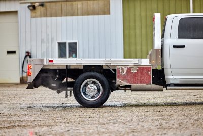 Side profile view of an EBY Big Country aluminum flat bed mounted on a white heavy-duty truck, showing the open flat deck, rear tail lights, EBY-branded side rail, chrome dual rear wheels, and a red under-bed storage box, parked on gravel in front of a metal farm building.